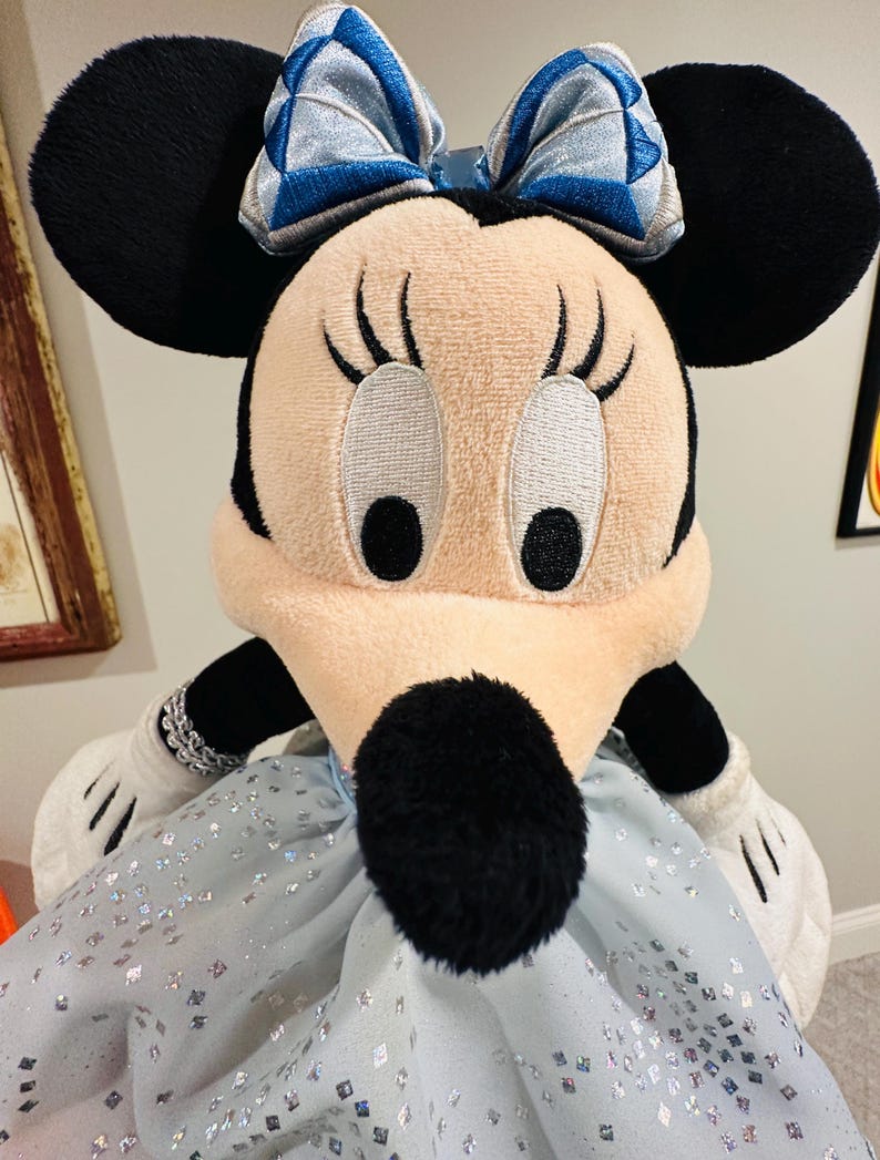 K&ouml;nnte beinhalten: Ein Pl&uuml;sch Minnie Mouse Puppe mit schwarzen Ohren, einem hellrosa Gesicht und einer schwarzen Nase. Sie tr&auml;gt ein hellblaues Kleid mit silbernen Glitzer und einer blau-silbernen Schleife. Ihre wei&szlig;en Handschuhe und schwarzen Arme sind sichtbar.