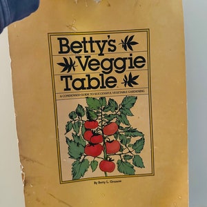 May include: A spiral-bound book titled "Betty's Veggie Table" with a yellow cover. The cover features a tomato plant illustration and the text "A Condensed Guide to Successful Vegetable Gardening" and "By Betty L. Grunow".