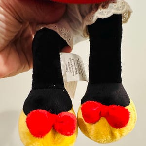Puede incluir: Un juguete de peluche con piernas negras y pies amarillos, cada uno adornado con un lazo rojo. El juguete tiene una parte superior roja y un vestido blanco con ribete de encaje. La etiqueta de Disney Store es visible.