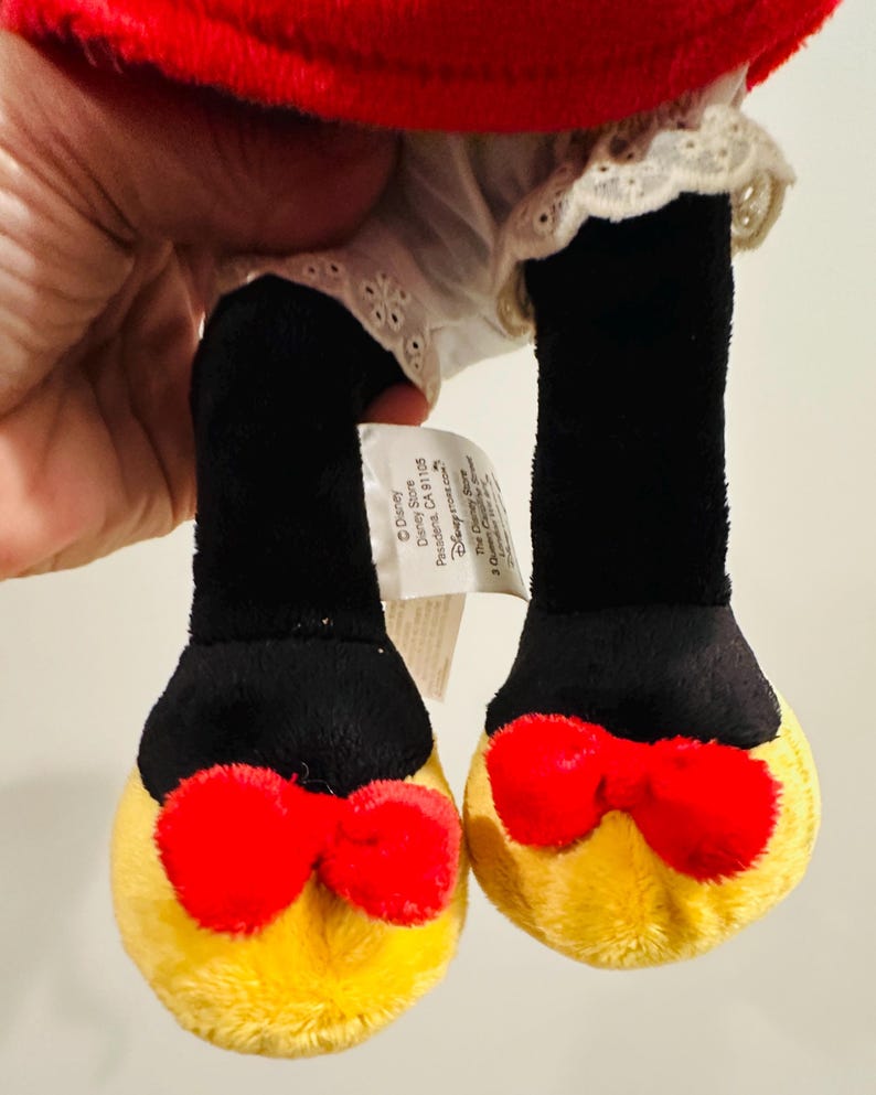 Puede incluir: Un juguete de peluche con piernas negras y pies amarillos, cada uno adornado con un lazo rojo. El juguete tiene una parte superior roja y un vestido blanco con ribete de encaje. La etiqueta de Disney Store es visible.