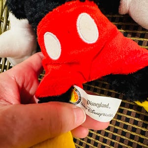 Puede incluir: Primer plano de un peluche de Mickey Mouse. El juguete presenta un guante rojo con &oacute;valos blancos, pelaje negro y una prenda amarilla. Una etiqueta dice "Disneyland, Walt Disney World."