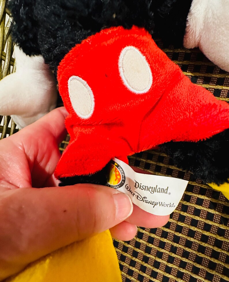 Puede incluir: Primer plano de un peluche de Mickey Mouse. El juguete presenta un guante rojo con &oacute;valos blancos, pelaje negro y una prenda amarilla. Una etiqueta dice "Disneyland, Walt Disney World."