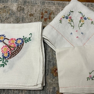 May include: White linen napkins and towels with colorful floral embroidery. One towel features a basket of flowers, while others have floral sprigs. The embroidery includes pink, blue, yellow, and green thread.