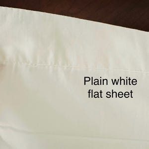 May include: A plain white flat sheet, with the text "Plain white flat sheet" in black. The sheet is made of a smooth, white fabric. The image shows a close-up of the sheet's edge.