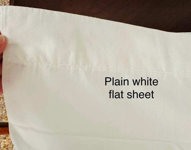 May include: A plain white flat sheet, with the text "Plain white flat sheet" in black. The sheet is made of a smooth, white fabric. The image shows a close-up of the sheet's edge.