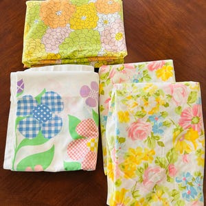 May include: Four folded vintage floral print bedsheets. One sheet features large flowers in yellow, orange, and green. Another has blue gingham flowers and pink and red checkered accents. The other two sheets have floral patterns in pink, yellow, and blue.