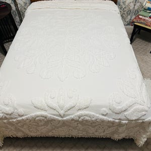 May include: A white bedspread with a raised floral pattern covers a bed. The bedspread has a scalloped edge and a textured design. The headboard is made of wood. The bed is in a room with patterned wallpaper.