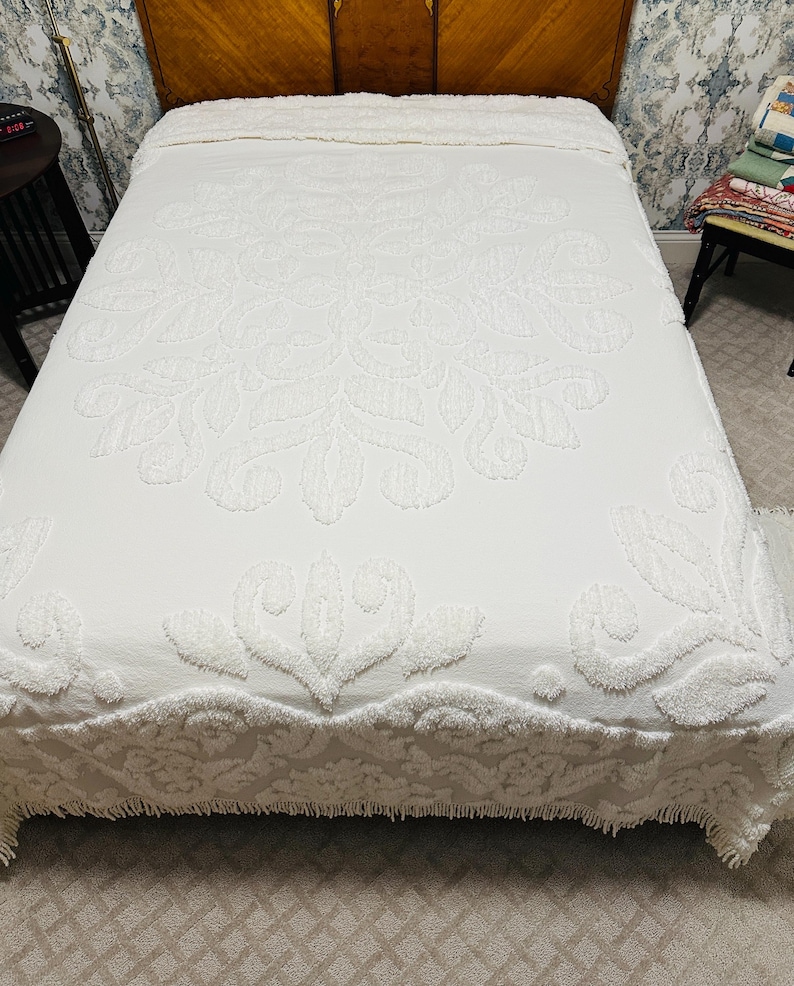 May include: A white bedspread with a raised floral pattern covers a bed. The bedspread has a scalloped edge and a textured design. The headboard is made of wood. The bed is in a room with patterned wallpaper.