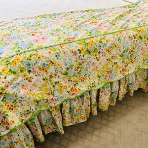 May include: A floral bedspread with a ruffled skirt. The fabric features a pattern of small flowers in shades of yellow, pink, blue, and green on a white background. A thin green trim outlines the edges of the bedspread.