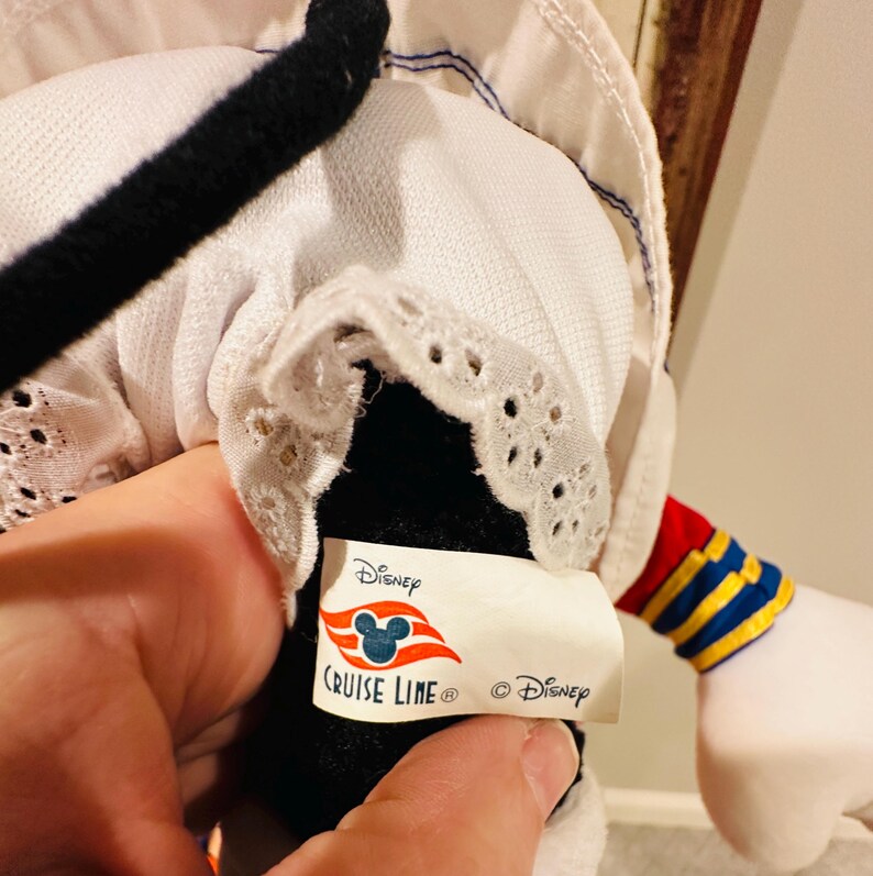 May include: A Disney Cruise Line plush toy featuring a black and white design with a white sailor hat and a red, yellow, and blue striped sleeve. The Disney Cruise Line logo is visible on a tag.