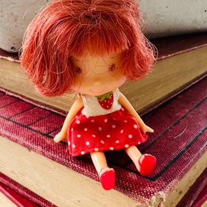 May include: A vintage Strawberry Shortcake doll with red hair, wearing a white top with a strawberry and a red polka-dot dress. The doll is sitting on a stack of old books with red covers.