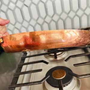 May include: A copper-colored, shallow pan with two handles. The pan is resting on a black gas stove. The pan has a decorative leaf pattern on the side. The pan is made of copper and has a shiny finish.