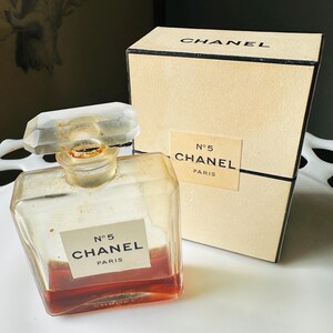 K&ouml;nnte beinhalten: Ein Vintage-Chanel-No. 5-Parf&uuml;mflakon mit einem klaren, facettierten Verschluss und einer rechteckigen Form. Die Flasche enth&auml;lt eine rotbraune Fl&uuml;ssigkeit und tr&auml;gt ein Etikett mit der Aufschrift "N&deg;5 CHANEL PARIS". Eine passende Schachtel befindet sich hinter der Flasche.