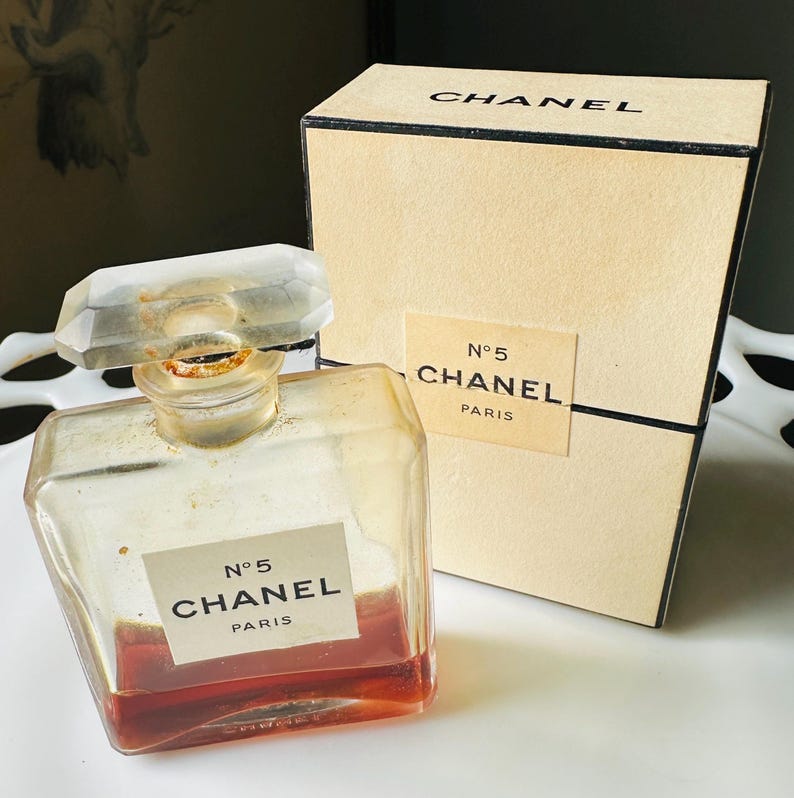 K&ouml;nnte beinhalten: Ein Vintage-Chanel-No. 5-Parf&uuml;mflakon mit einem klaren, facettierten Verschluss und einer rechteckigen Form. Die Flasche enth&auml;lt eine rotbraune Fl&uuml;ssigkeit und tr&auml;gt ein Etikett mit der Aufschrift "N&deg;5 CHANEL PARIS". Eine passende Schachtel befindet sich hinter der Flasche.