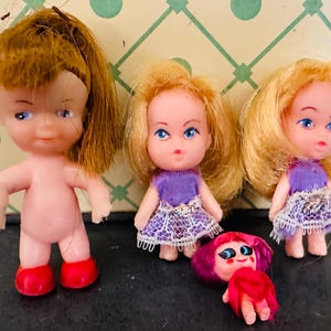 May include: A collection of vintage plastic dolls. One doll is nude with red boots, and the others wear purple dresses with lace skirts. One doll's head and arms are detached. The dolls have blonde and brown hair.