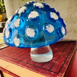 May include: A glass mushroom figurine featuring a translucent blue cap adorned with raised, opaque white circular spots. The mushroom has a clear, solid glass stem that supports the rounded cap, creating a smooth and reflective surface throughout.
