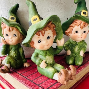 May include: Three ceramic figurines of leprechauns wearing green hats with yellow buckles and green outfits. The figurines have rosy cheeks, brown hair, and are sitting on a red book.