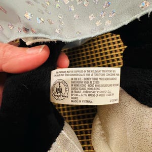 K&ouml;nnte beinhalten: Ein schwarz-wei&szlig;es Pl&uuml;schtier mit silbernem Paillettenstoff und hellblauem, glitzerndem Stoff. Das Spielzeug hat ein Etikett mit Text, darunter "Disney Parks" und "Made in Vietnam."