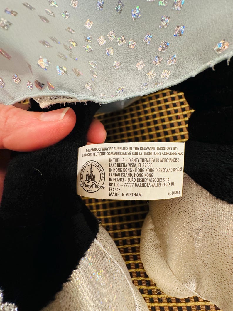 K&ouml;nnte beinhalten: Ein schwarz-wei&szlig;es Pl&uuml;schtier mit silbernem Paillettenstoff und hellblauem, glitzerndem Stoff. Das Spielzeug hat ein Etikett mit Text, darunter "Disney Parks" und "Made in Vietnam."