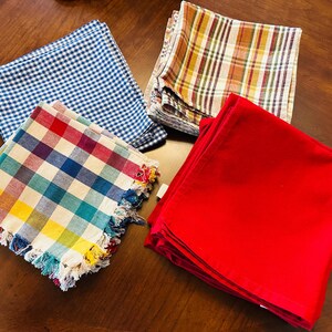 May include: A collection of folded cloth napkins in various patterns and colors. Includes a blue and white gingham, a plaid with red, yellow, and brown, a red solid, and a colorful plaid with fringe.