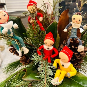 May include: A festive arrangement featuring vintage elf ornaments in various colors, including red, blue, and yellow. The elves are posed among pine cones, greenery, and large green leaves, creating a whimsical holiday display.
