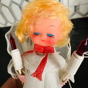May include: A vintage Christmas angel doll with blonde hair, blue eyes, and rosy cheeks. The doll wears a white dress with a red bow and holds two red and white Christmas lights. A delicate white lace halo frames the doll's face.
