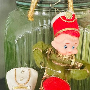 May include: Three elf figurines with painted faces and felt clothing. One elf wears red and gold, another wears green and red with a drum, and the third wears white and red with a drum. They are positioned in front of a large green glass jar.