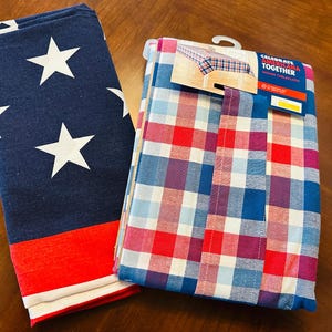 May include: Two folded tablecloths. One is navy blue with white stars and a red stripe. The other is a woven plaid in red, blue, and purple. The tag reads "Celebrate Americana Together Woven Tablecloth".