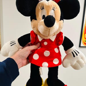 May include: A plush Minnie Mouse doll, featuring black ears, a red and white polka dot dress, and yellow shoes with red bows. The doll has white gloves and a red bow on her head.