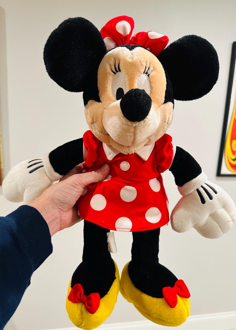May include: A plush Minnie Mouse doll, featuring black ears, a red and white polka dot dress, and yellow shoes with red bows. The doll has white gloves and a red bow on her head.