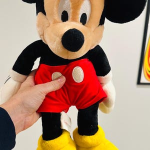 May include: A plush Mickey Mouse doll, featuring black ears, a tan face, and a black nose. He wears red shorts with white buttons, black arms, white gloves, and yellow shoes. The doll is approximately 18 inches tall.
