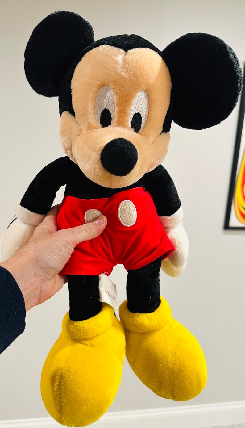 May include: A plush Mickey Mouse doll, featuring black ears, a tan face, and a black nose. He wears red shorts with white buttons, black arms, white gloves, and yellow shoes. The doll is approximately 18 inches tall.