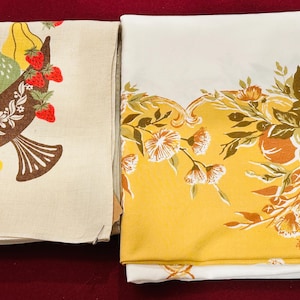 May include: Two folded rectangular fabric cloths. The left cloth is beige with a brown cornucopia filled with yellow, green, and red fruit. The right cloth is white with a wide yellow border featuring a printed design of orange and green fruit and flowers.