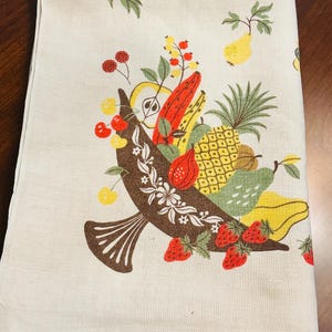 May include: A cream-colored kitchen towel featuring a brown cornucopia overflowing with a colorful assortment of fruits including a yellow pineapple, red strawberries, cherries, and various other stylized fruits in shades of yellow, red, and green.