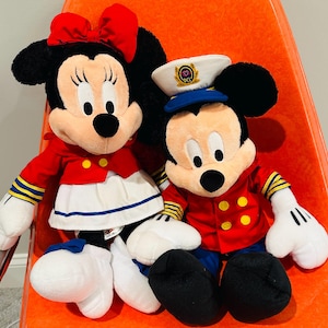 May include: Two plush Disney character dolls, Minnie Mouse and Mickey Mouse, are seated together. Minnie wears a red bow, jacket, and skirt. Mickey wears a captain's hat and red jacket. Both have black ears and white gloves.