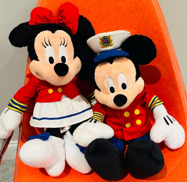 May include: Two plush Disney character dolls, Minnie Mouse and Mickey Mouse, are seated together. Minnie wears a red bow, jacket, and skirt. Mickey wears a captain's hat and red jacket. Both have black ears and white gloves.