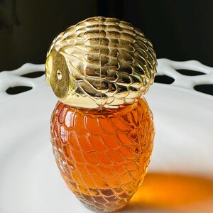 K&ouml;nnte beinhalten: Eine dekorative Parf&uuml;mflasche mit einem goldfarbenen, eulenartigen Verschluss und einem strukturierten, bernsteinfarbenen Glask&ouml;rper. Die Flasche steht auf einem wei&szlig;en, verzierten Teller. Das Design der Flasche erinnert an Vintage-Parf&uuml;mflaschen.