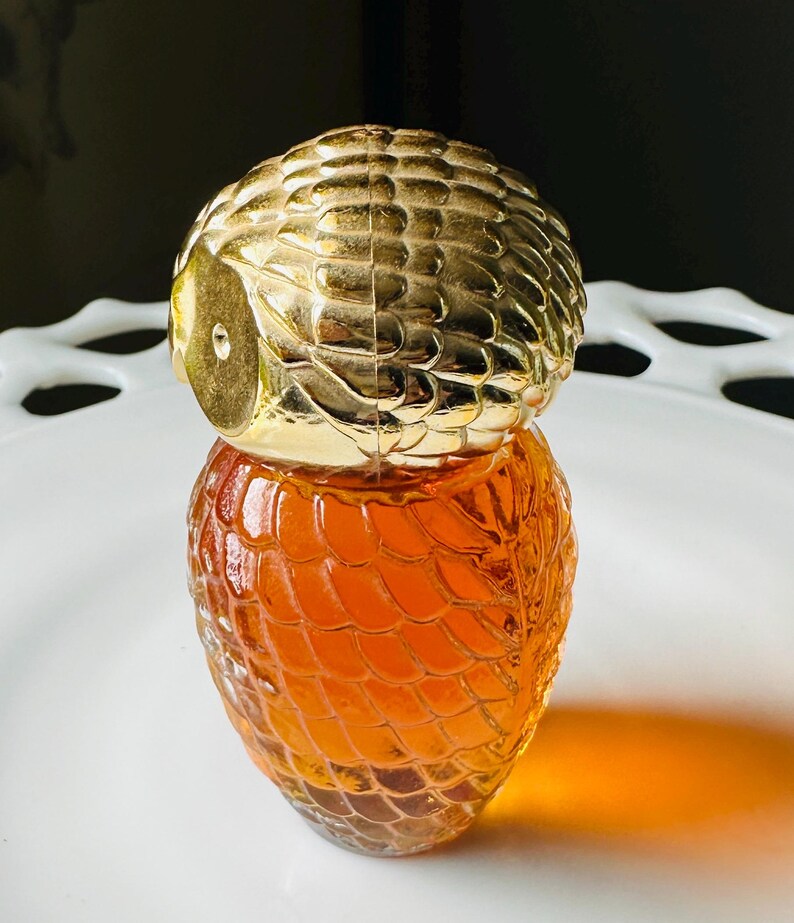 K&ouml;nnte beinhalten: Eine dekorative Parf&uuml;mflasche mit einem goldfarbenen, eulenartigen Verschluss und einem strukturierten, bernsteinfarbenen Glask&ouml;rper. Die Flasche steht auf einem wei&szlig;en, verzierten Teller. Das Design der Flasche erinnert an Vintage-Parf&uuml;mflaschen.