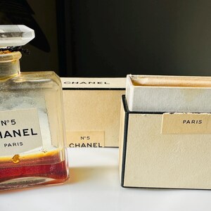 K&ouml;nnte beinhalten: Eine Vintage-Parf&uuml;mflasche von Chanel No. 5 mit einem klaren, facettierten Verschluss und einem Etikett mit der Aufschrift "N&deg;5 CHANEL PARIS". Die Flasche enth&auml;lt eine rotbraune Fl&uuml;ssigkeit. Zwei beige Schachteln mit schwarzem Rand sind ebenfalls vorhanden, eine mit der Aufschrift "N&deg;5 CHANEL" und die andere "PARIS".