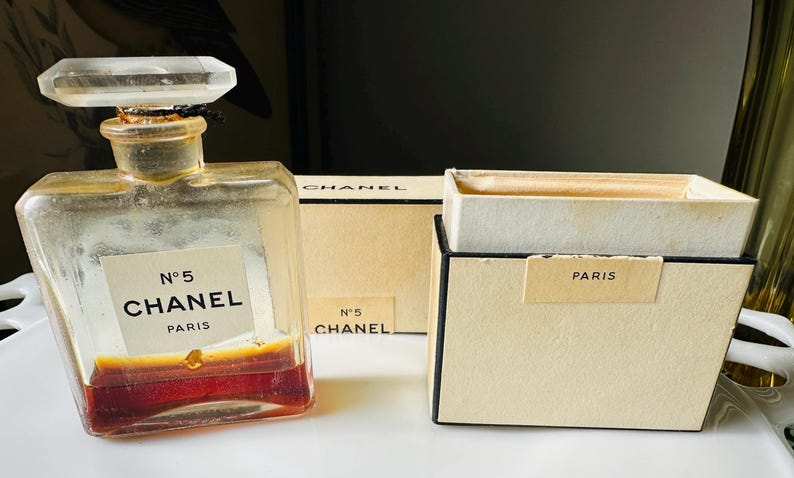 K&ouml;nnte beinhalten: Eine Vintage-Parf&uuml;mflasche von Chanel No. 5 mit einem klaren, facettierten Verschluss und einem Etikett mit der Aufschrift "N&deg;5 CHANEL PARIS". Die Flasche enth&auml;lt eine rotbraune Fl&uuml;ssigkeit. Zwei beige Schachteln mit schwarzem Rand sind ebenfalls vorhanden, eine mit der Aufschrift "N&deg;5 CHANEL" und die andere "PARIS".