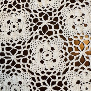 May include: Close-up of a white, intricately crocheted doily. The design features repeating floral and geometric patterns, creating a delicate and textured surface. The doily is set against a dark brown background, highlighting the detailed craftsmanship.