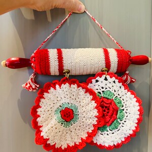 May include: A decorative kitchen set featuring a crochet rolling pin with red and white stripes and tassels, along with two matching crochet potholders. The potholders have a floral design with red, green, and white accents. The set is designed for hanging.