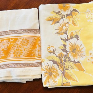 May include: Two vintage textiles: a folded white towel with an orange floral and brown geometric border, and a yellow tablecloth with a large floral design in white, yellow, and brown. The tablecloth has a white background with a yellow border.