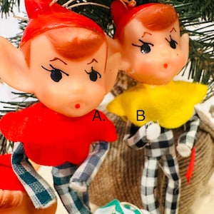 May include: Vintage Christmas elf ornaments. Two elf ornaments with red hats and faces. One elf wears a red shirt and blue and white plaid pants. The other elf wears a yellow collar and black and white plaid pants. The ornaments are attached to a Christmas tree.