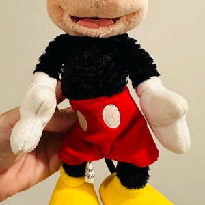 Puede incluir: Un peluche de Mickey Mouse. El juguete presenta un cuerpo, orejas y cola negros, con una cara beige y guantes blancos. Mickey lleva pantalones cortos rojos con botones blancos y zapatos amarillos. Un personaje cl&aacute;sico de Disney.
