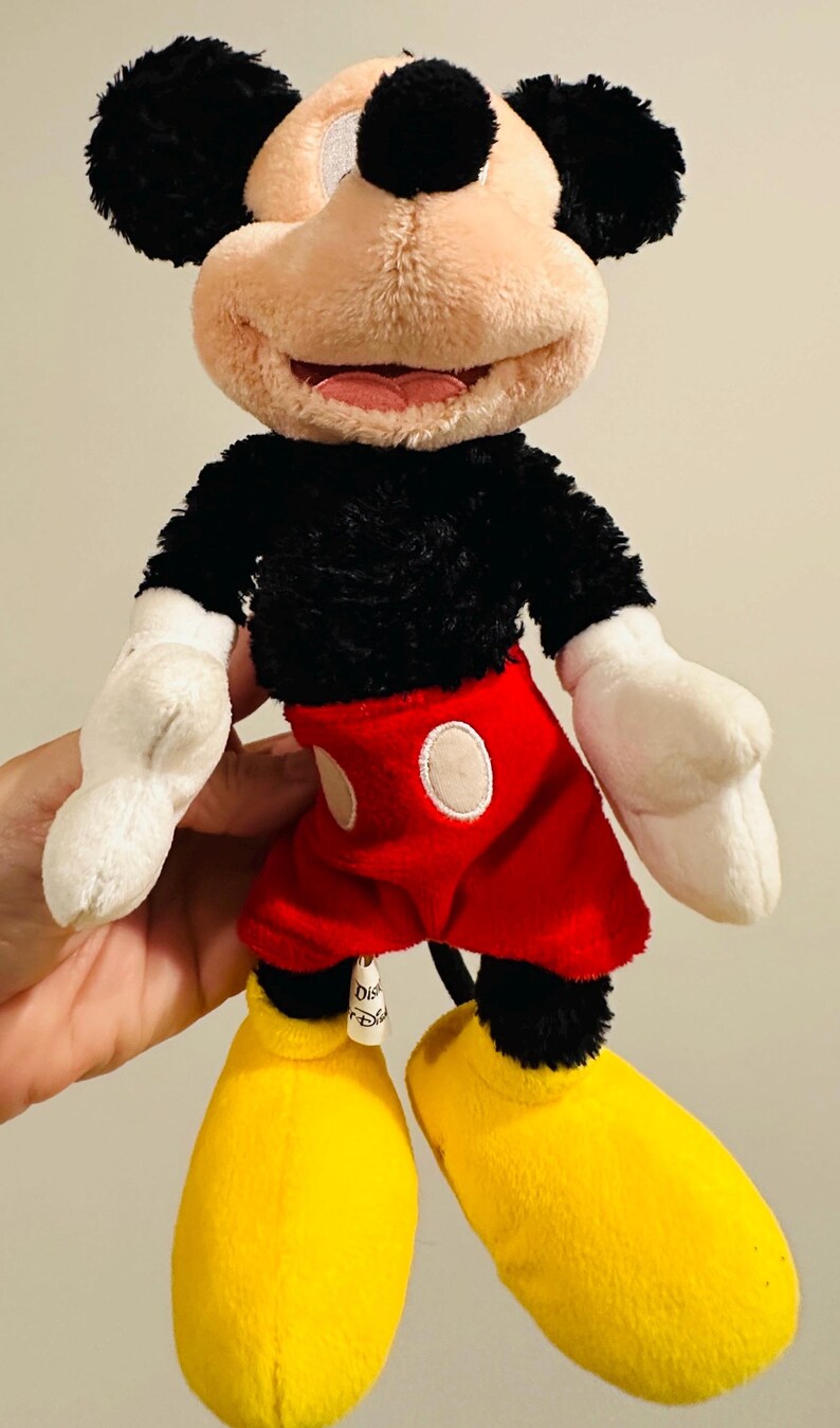 Puede incluir: Un peluche de Mickey Mouse. El juguete presenta un cuerpo, orejas y cola negros, con una cara beige y guantes blancos. Mickey lleva pantalones cortos rojos con botones blancos y zapatos amarillos. Un personaje cl&aacute;sico de Disney.