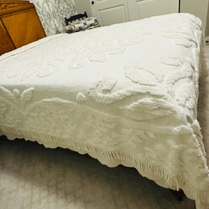 May include: A white bedspread with a raised floral pattern and a fringed edge. The bedspread is covering a bed with a wooden headboard. The room has a neutral color palette.