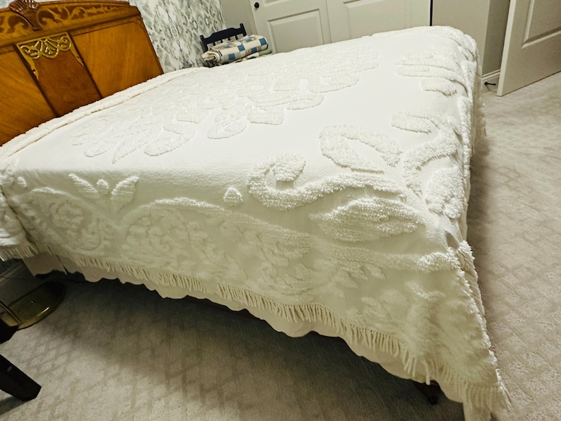 May include: A white bedspread with a raised floral pattern and a fringed edge. The bedspread is covering a bed with a wooden headboard. The room has a neutral color palette.