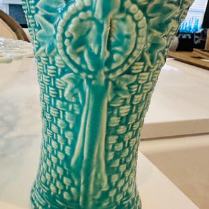May include: A turquoise ceramic vase with a basket weave design. The vase features a central vertical element with a leaf and circle motif. The vase is approximately 10 inches tall.
