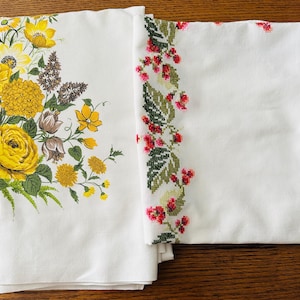 May include: Two white tablecloths. One features a large floral design with yellow, brown, and green flowers. The other has a cross-stitched border of red berries and green leaves. Both are folded and displayed on a wooden surface.